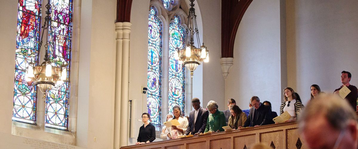 Members of the congregation stand to sing in the balcony. Stained glass and the bright chapel chandeliers over look the worshippers. The photo is taken from the floor, angled upwards.