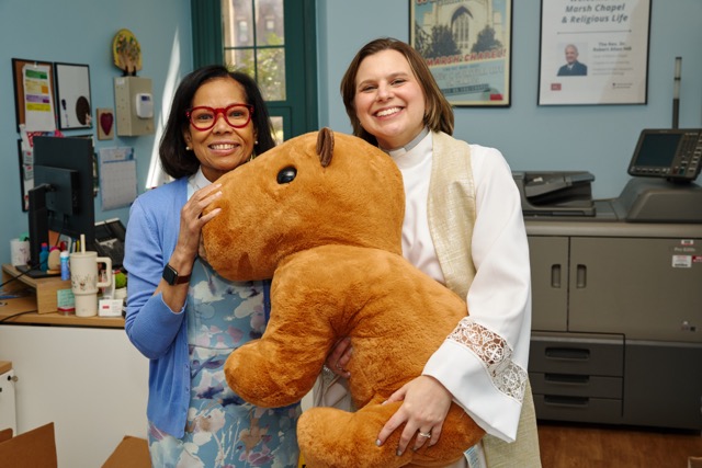 Rev. Karen and Rev. Jessica smile at the camera in the Marsh Chapel office while holding a large capybara stuffed animal