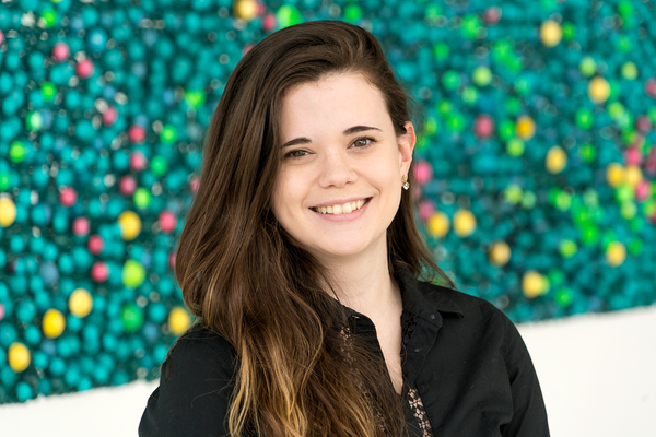Marie Vearling's headshot. Photo of a smiling person from their shoulders up. She has long brown hair, peach/white skin and is wearing a long-sleeved black buttoned-down shirt. The background behind them is speckled green dots with other accent colors.