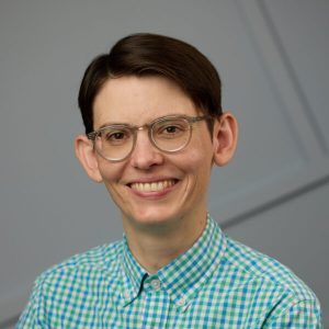 Allison Reilly's headshot. Photo of a smiling person from their shoulders up. They have short brown hair, clear-rimmed glasses, peach/white skin and are wearing a green and blue checkered shirt. The background behind them is gray with intersecting lines.
