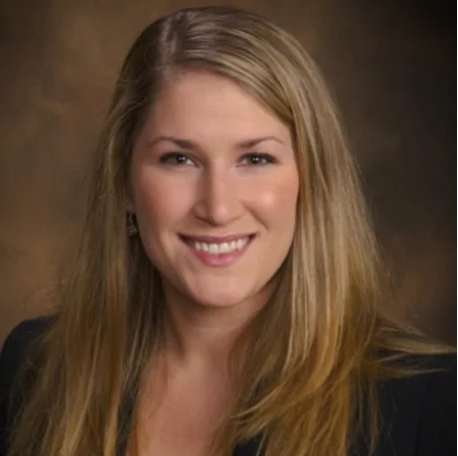 smiling female with light brown shoulder length straight hair, wearing dark suit jacket, background is marbled light brown and dark brown