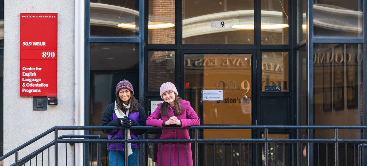 Two young women posing outside in winter hats and coats while leaning against a railing.