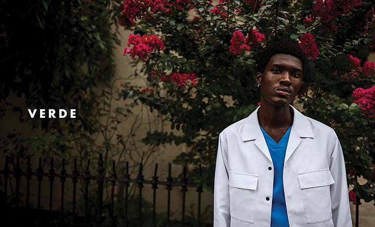 Photo: A young dark-skinned Black man models blue scrubs and a white lab coat in front of a large rose bush. The name 'VERDE' is shown to the left of him in bold, white letters.