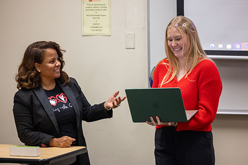 Two women stand at the front of a classroom in conversation with one another. One (at right) is holding a laptop.