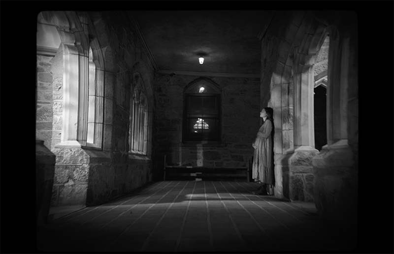 Production still from Forgive Me Father, showing a pregnant woman standing alone in a dimly lit hallway.