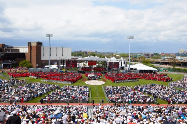 Distance view from the grandstand to the stage on Nickerson Field, filled with guests and graduates in regalia.