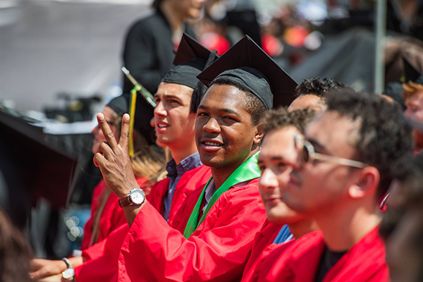 In a row of graduates, one turns to the camera and hold up the Peace sign with two fingers.