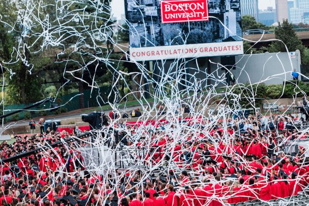 White paper streamers chaotically overhead a large group of BU graduates in red gowns with Congratulations Graduates on the jumbotron.