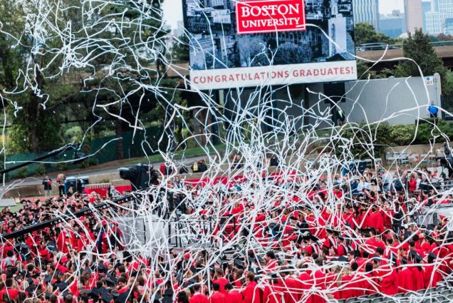 White paper streamers chaotically ovehead a large group of BU graduates in red gowns with Congratulations Graduates on the jumbotron.