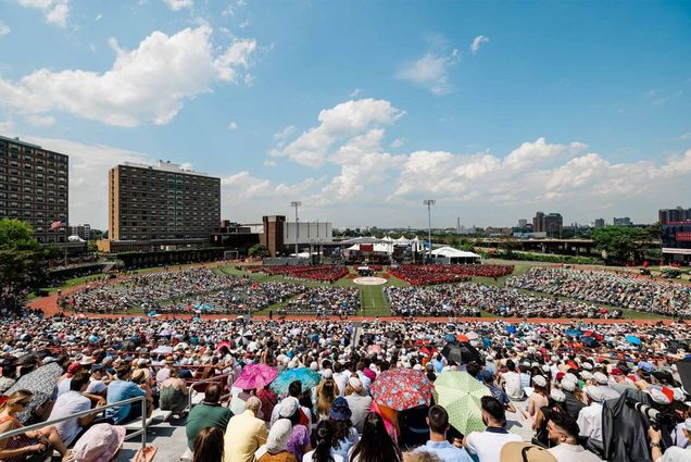 Photo of commencement attendees taken from the bleachers looking towards the stage. The backs of thousands of attendees are seen as well as the green turf of nickerson field. The sky is bright blue with a few small clouds.