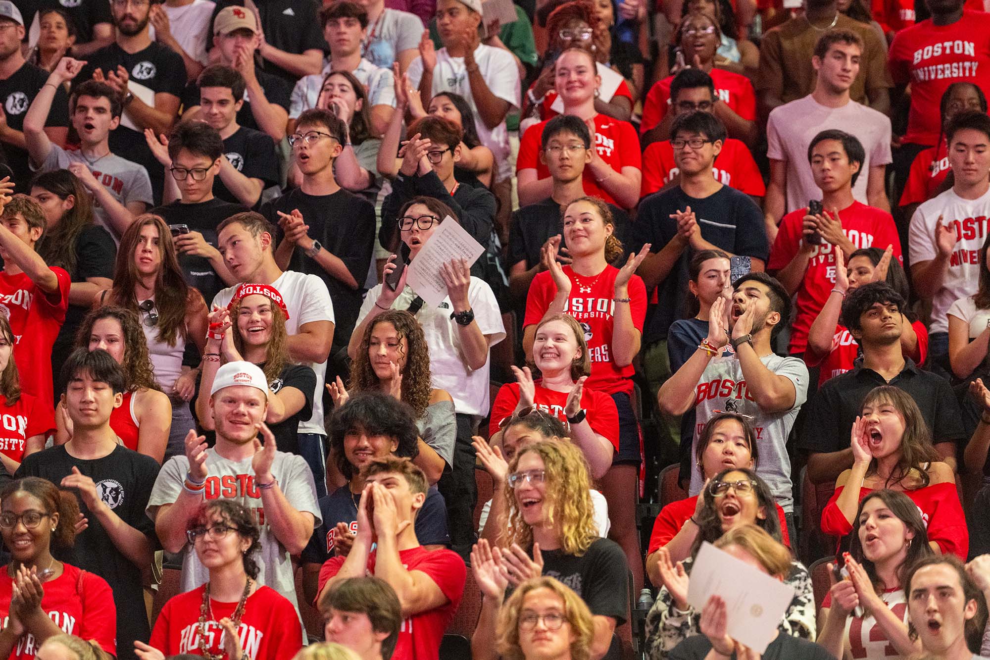 Crowd of students in the stands cheering, many wearing Boston University t-shirts in red, white, black, or grey.