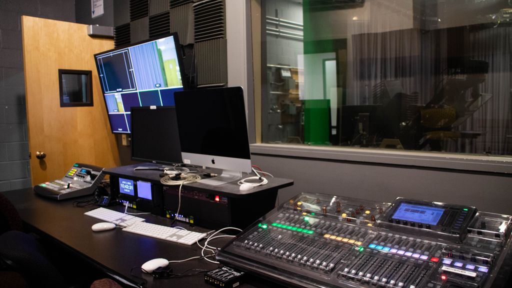 Decorative image that displays the control room of Studio East. Included is a control board, several screens, and a large audio mixer.