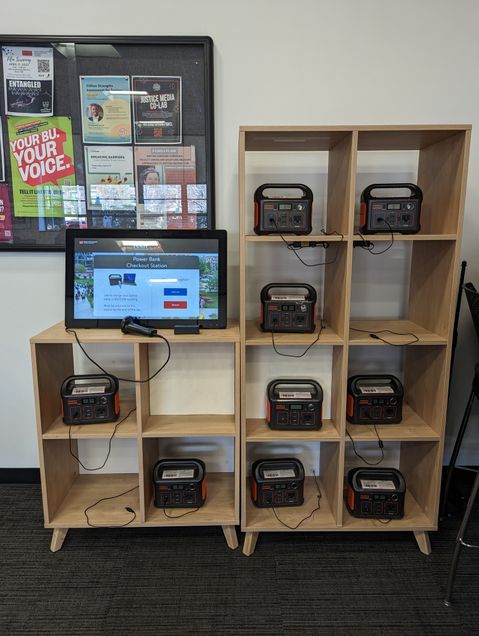 Image of a wall shelf full of Jackery Brand Power Banks located in the COM Student Lounge