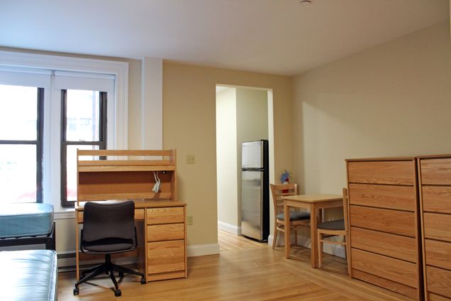 student apartment with desk, beds, dressers, table/chairs, and refrigerator.