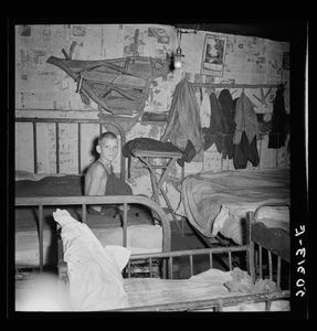 On the right side of the image a thin boy sits on a bed. The space around him is cluttered with three iron bedframes and clothing hanging from the walls, which are covered in what look to be newspaper ads. from