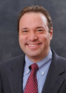 formal headshot of a man in a gray suit with a blue shirt and red tie, against a gray background