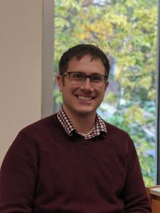 man with short brown hair and glasses, wearing a red sweater with a colloared. shirt underneath