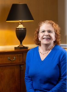 woman with short red hair in a blue shirt sitting in front of a bureau with a lamp on top of it.