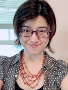Picture of a woman wearing glasses with a collared black polka dot shirt and an orange necklace