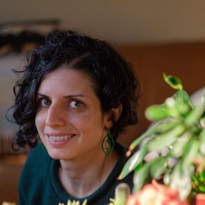 Woman standing by a plant. Women has black hair and a emerald green top.