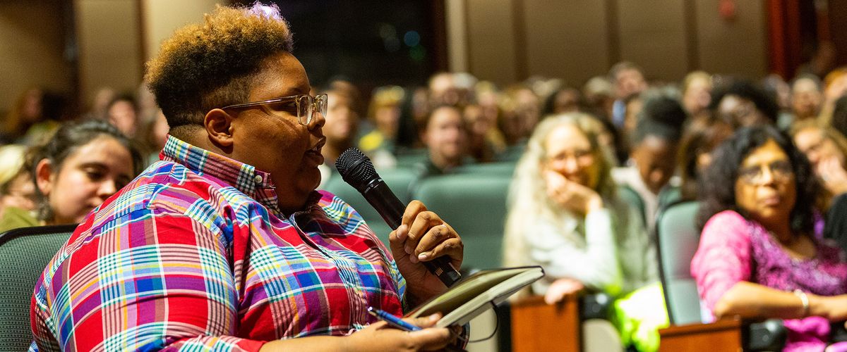 Black male speaking on a microphone in front of an auditorium full of colleagues