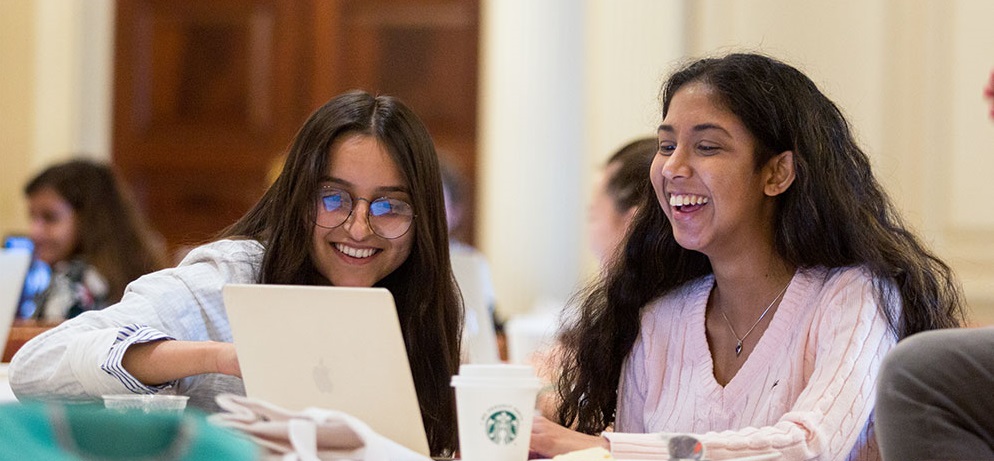 Two women seated at a table laughing and smiling behind a laptop screen