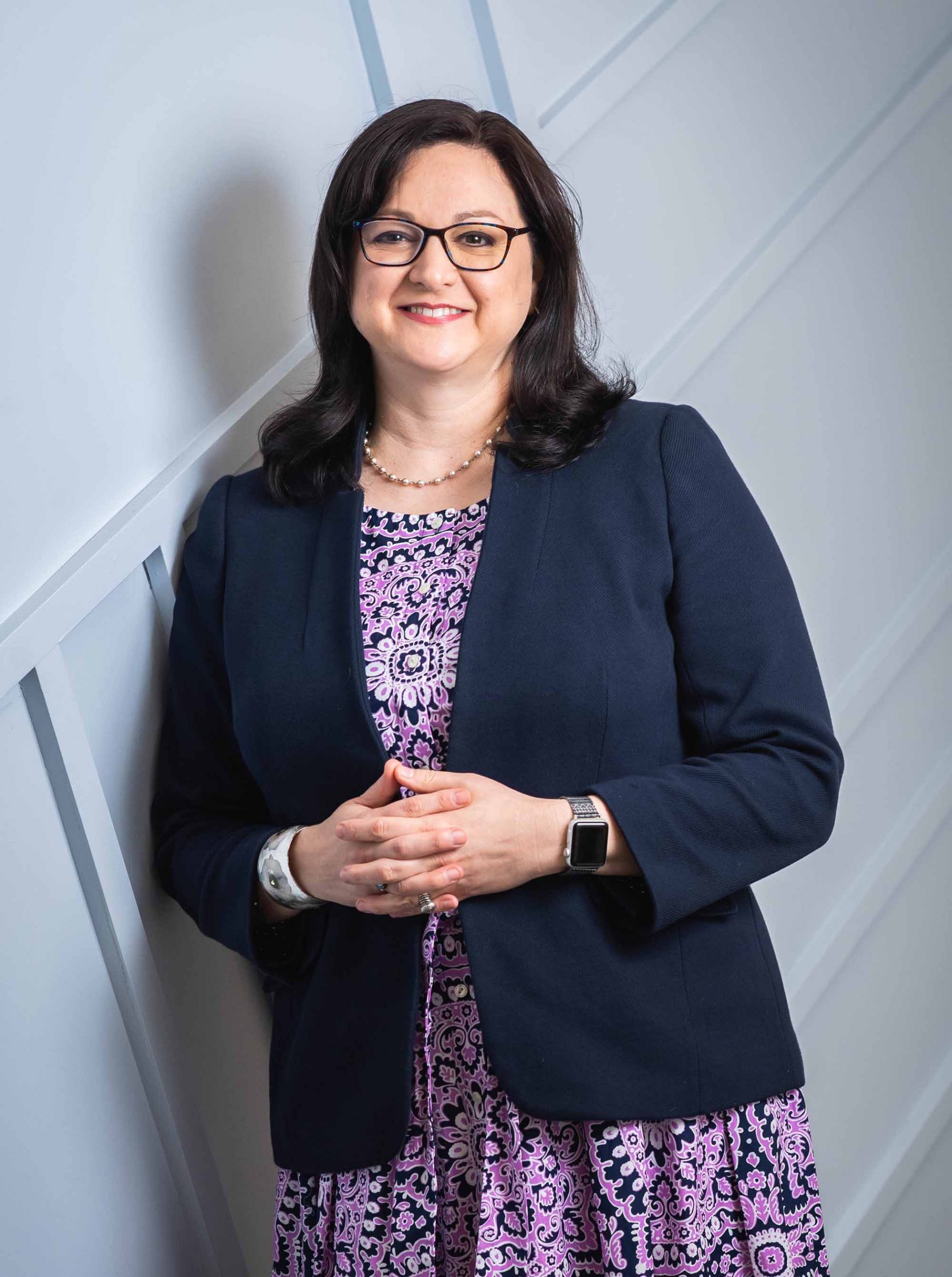 Debbie Bazarsky wearing glasses purple dress and dark blazer, leaning against a light blue wall and smiling.