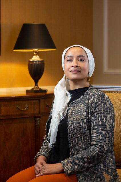 Photo of Karin Firoza. They are earing a white headscarf, orange pants, a black shirt, and a dark patterned jacket.