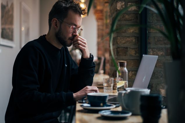 White man on a Apple laptop inside a coffeeshop next to coffee and water