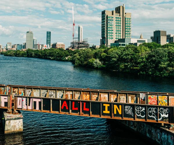 Photo of the Charles River and Boston skyline. Bridge in foreground has spray-painted words "ALL IN"