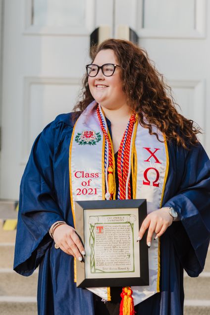 Woman in graduation regalia with stole holding a Chi Omega Sorority framed photo