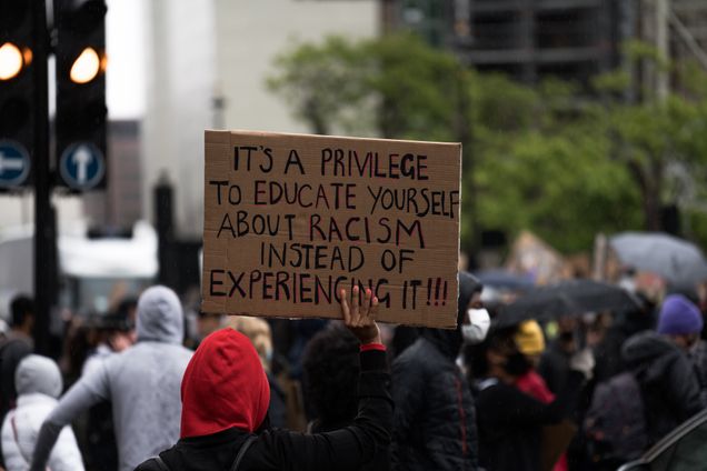 People gathered at a protests, one holding a sign stating: It's a privilege to educate yourself about racism instead of experiencing it!!!