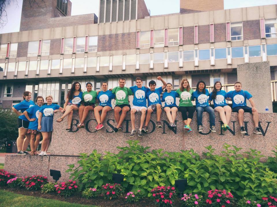 Students sit on a stone Boston University sign while wearing blue FYSOP shirts.