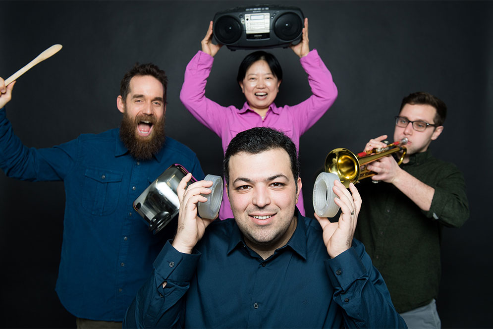 Boston University Ph. D. candidate Reza Ghaffarivardavagh poses with his research team. Reza holds the noise cancellation devices his team developed over his ears while his teammates make noise banging a pot, playing a trumpet, and a boombox in the background