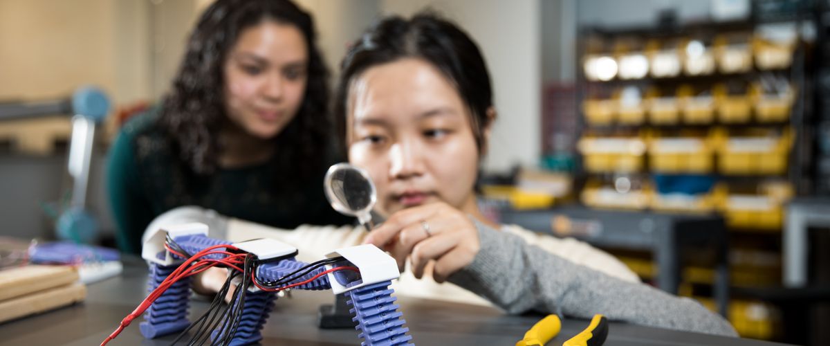 Student examining robotics work with a magnifying glass while a second student looks over her shoulder
