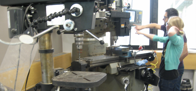 two students working at the nc mill in silab, pointing at the digital readout screen, with a drill press in the foreground