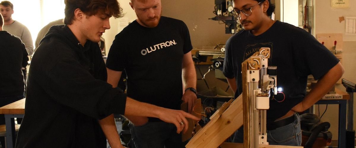 2 students with their lutron project and one of the judges; they are gesturing at the details on the back of their vertical automated light drawing board