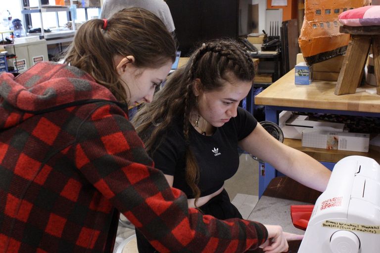 a student worker in the foreground leaning down to help a student in the midground with the sewing machine