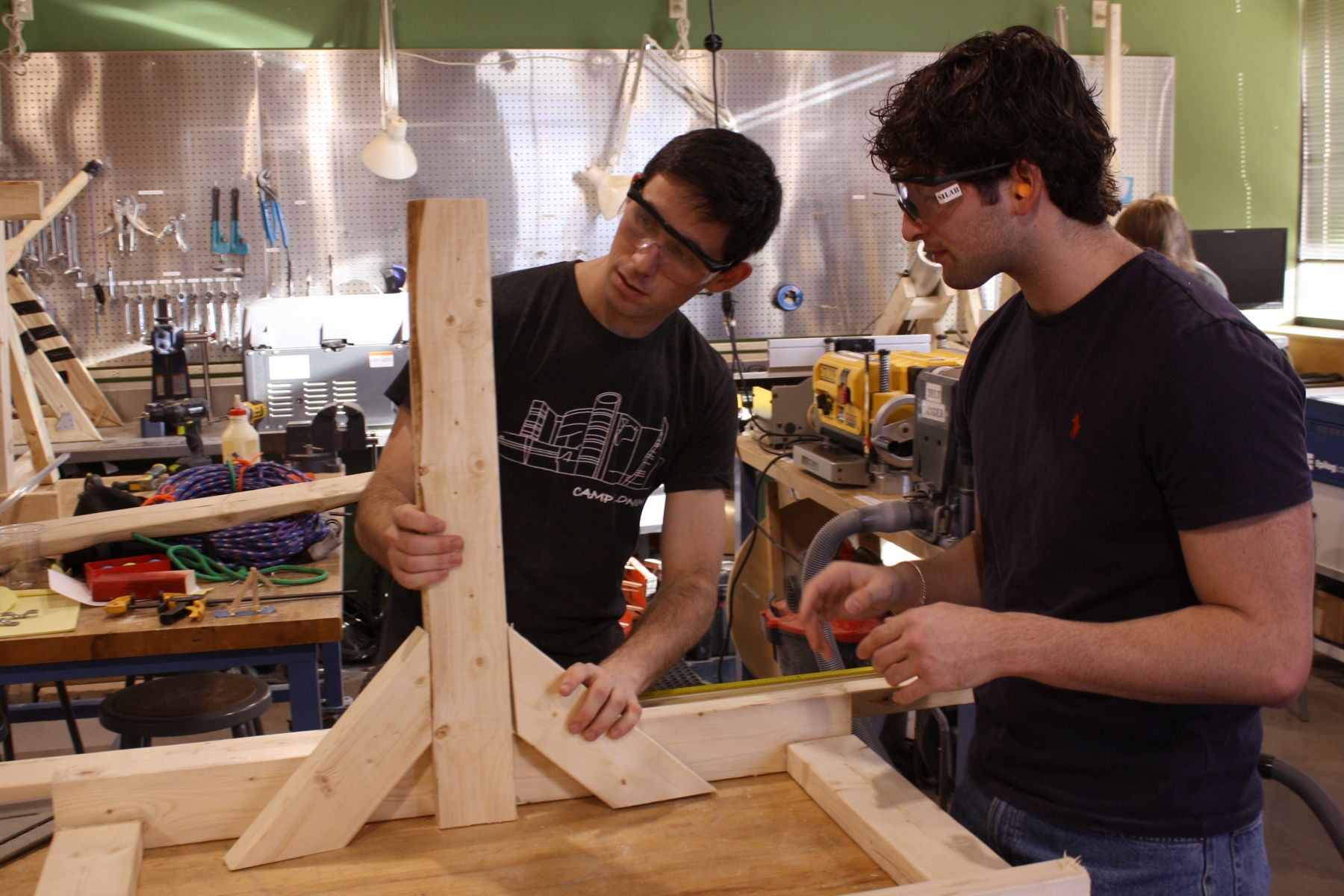 1 student holds the side support of a catapult against the wooden base frame while the other watches as the two discuss