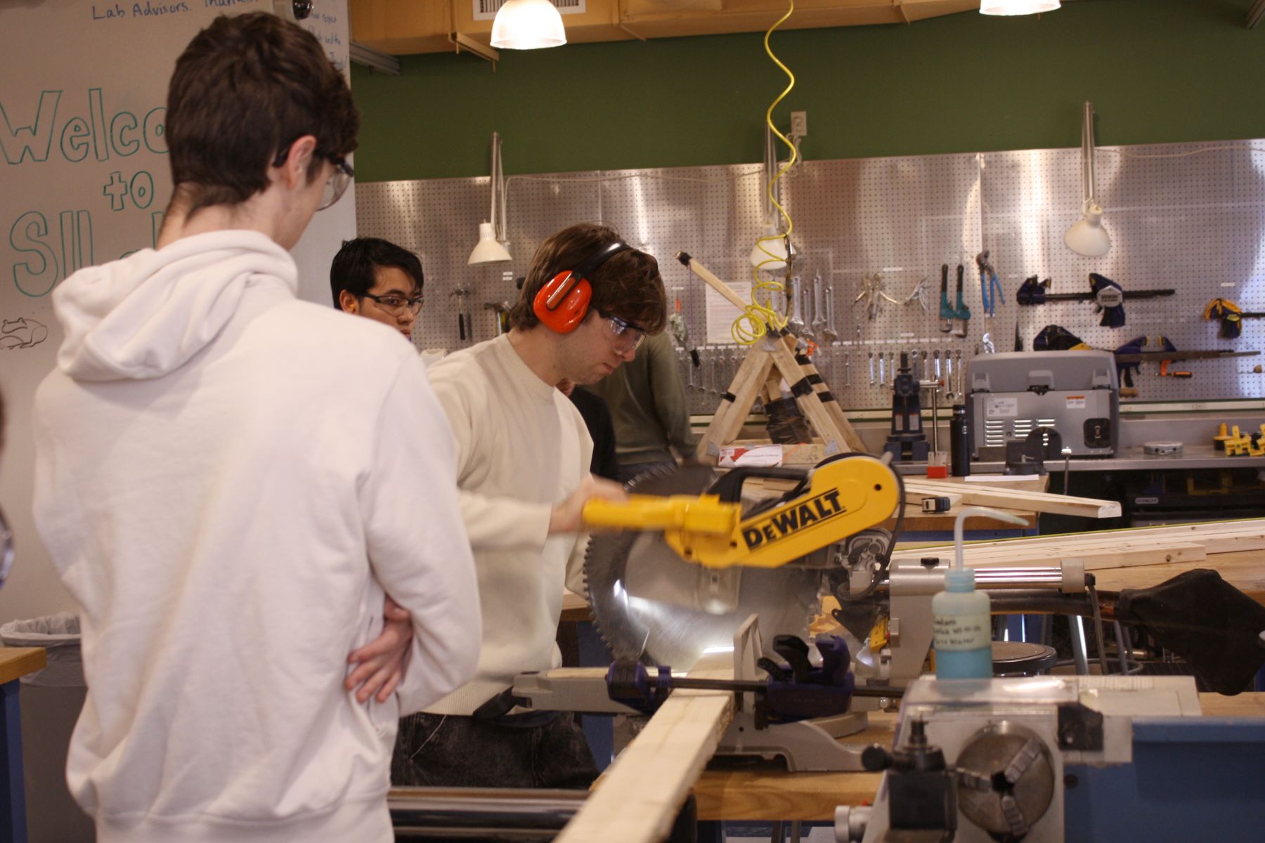 a student facing away from the camera watches as his teammate cuts a 2x4 board on the miter saw