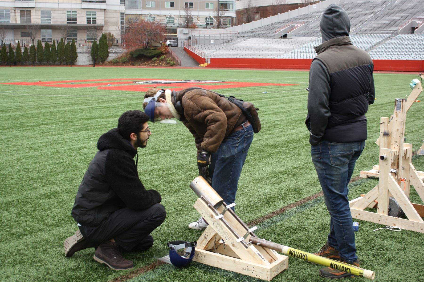 2 students crouched and working on their catapult while another looks downfield at the competition