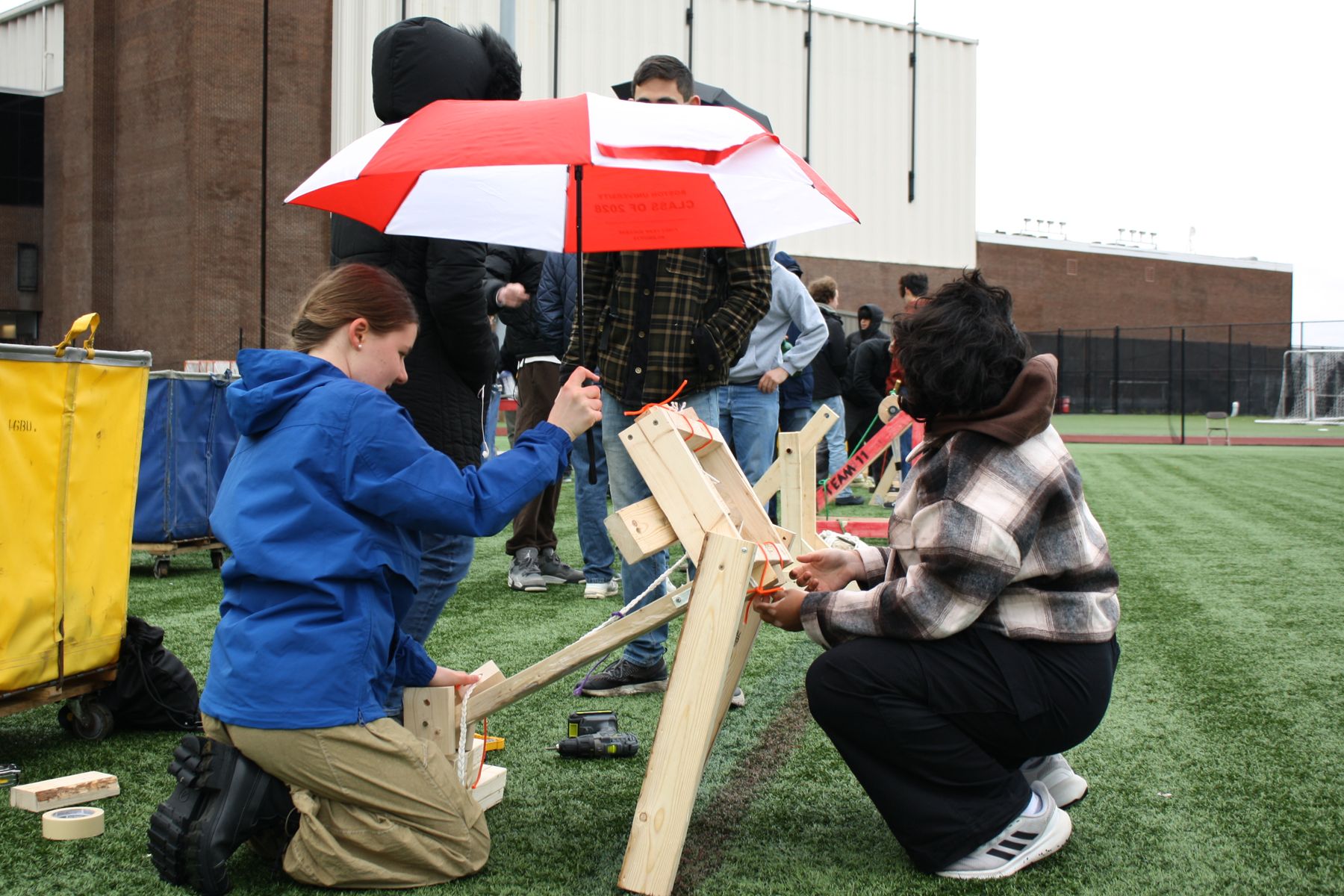 2 students crouch and work on their catapult while holding a BU umbrella against the mist