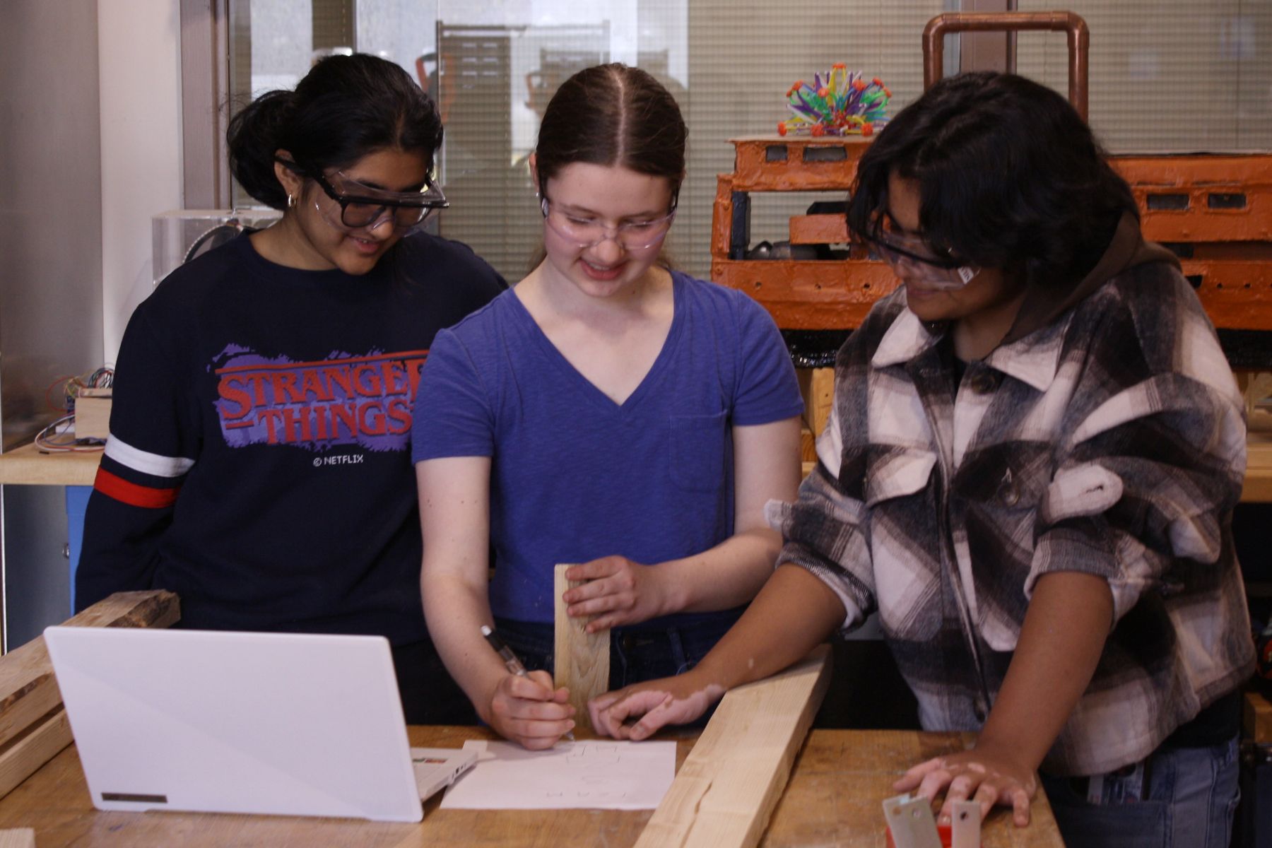 3 students standing around as they sketch plans for their catapult