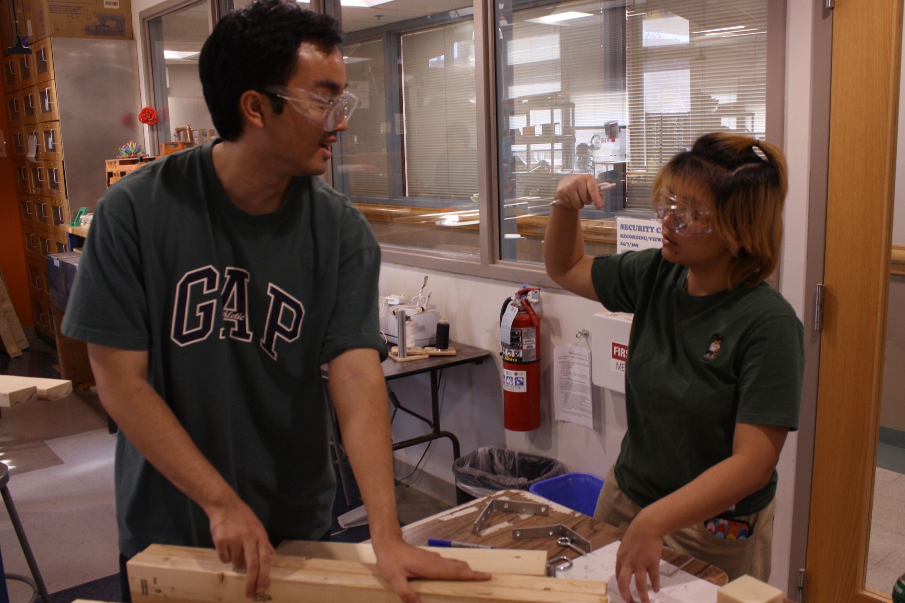 2 students in green shirts holding boards and gesturing with their hands what they plan to cut