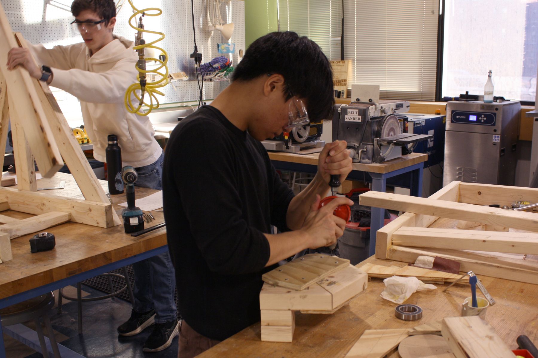 a student in the foreground manually drilling a tiny hole in a board, a student in the background is holding the frame of his catapult in place