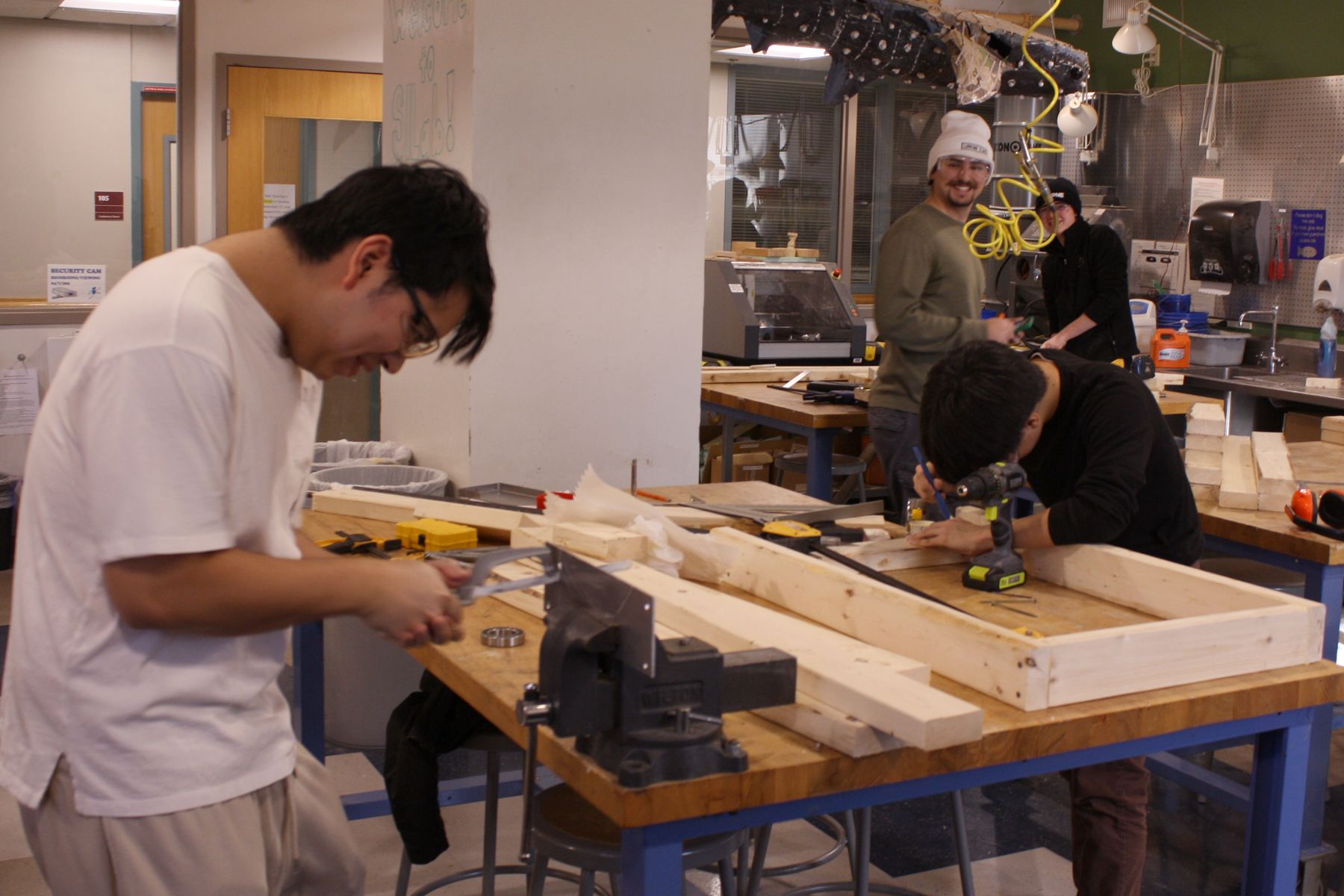 2 students in the foreground workng on cutting a piece of aluminum and marking wood, 2 students in the background are turned to look at the 2