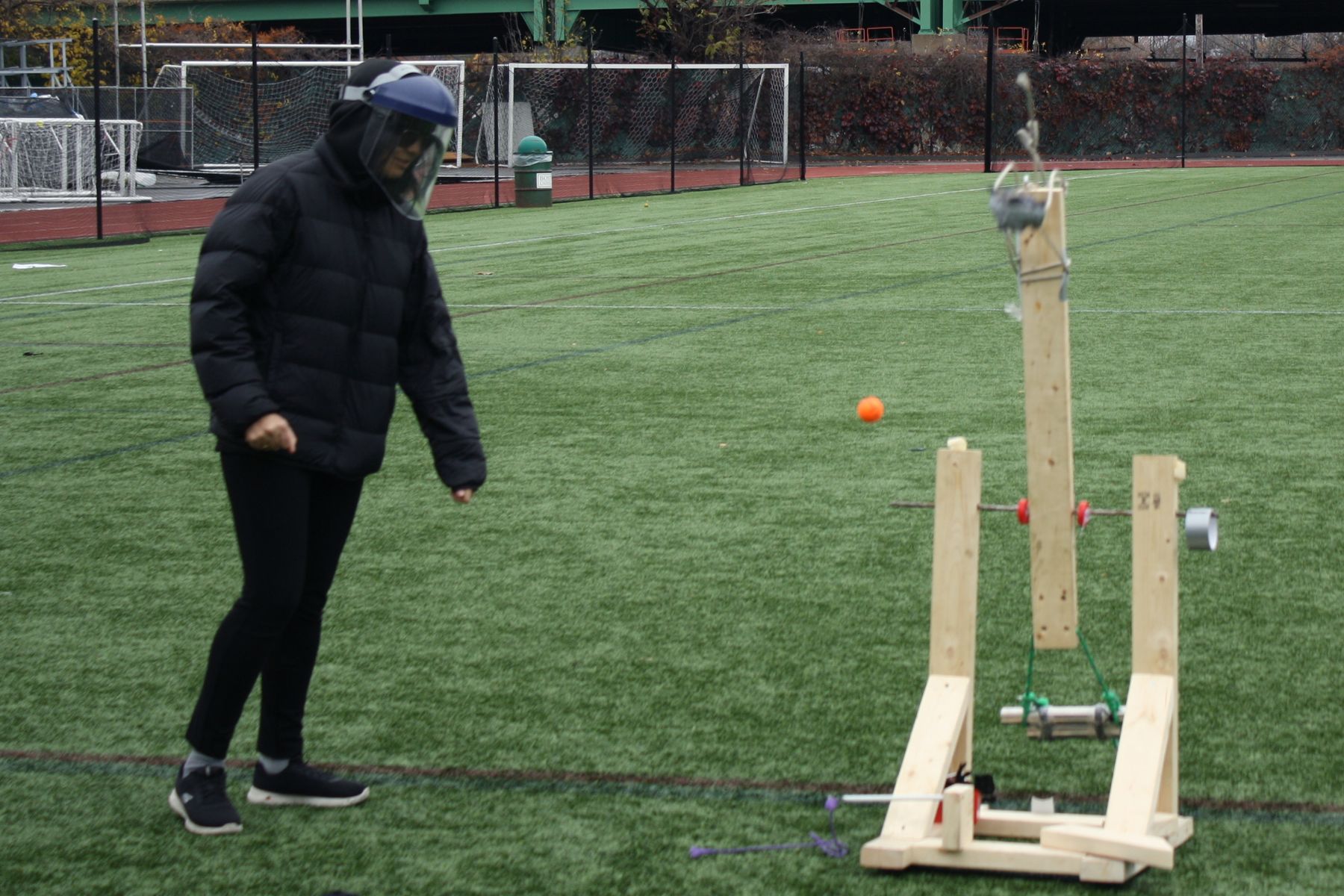 a student stands at a distance while launching her catapult, the orange golf ball is caught in motion as it starts to launch