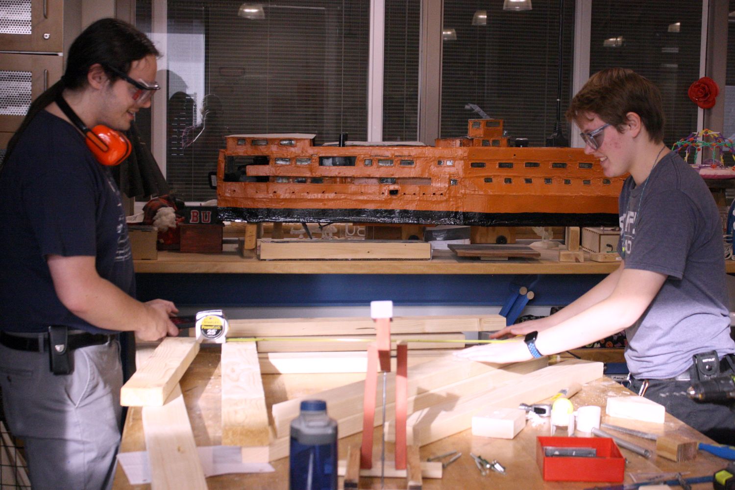 2 students measuring their boards as they work on building their catapult to match the model they made that is on the table in the foreground