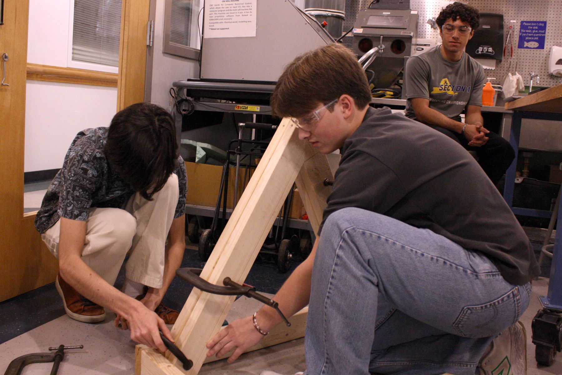 2 students crouched over their catapult clamping parts together while their teammate looks on