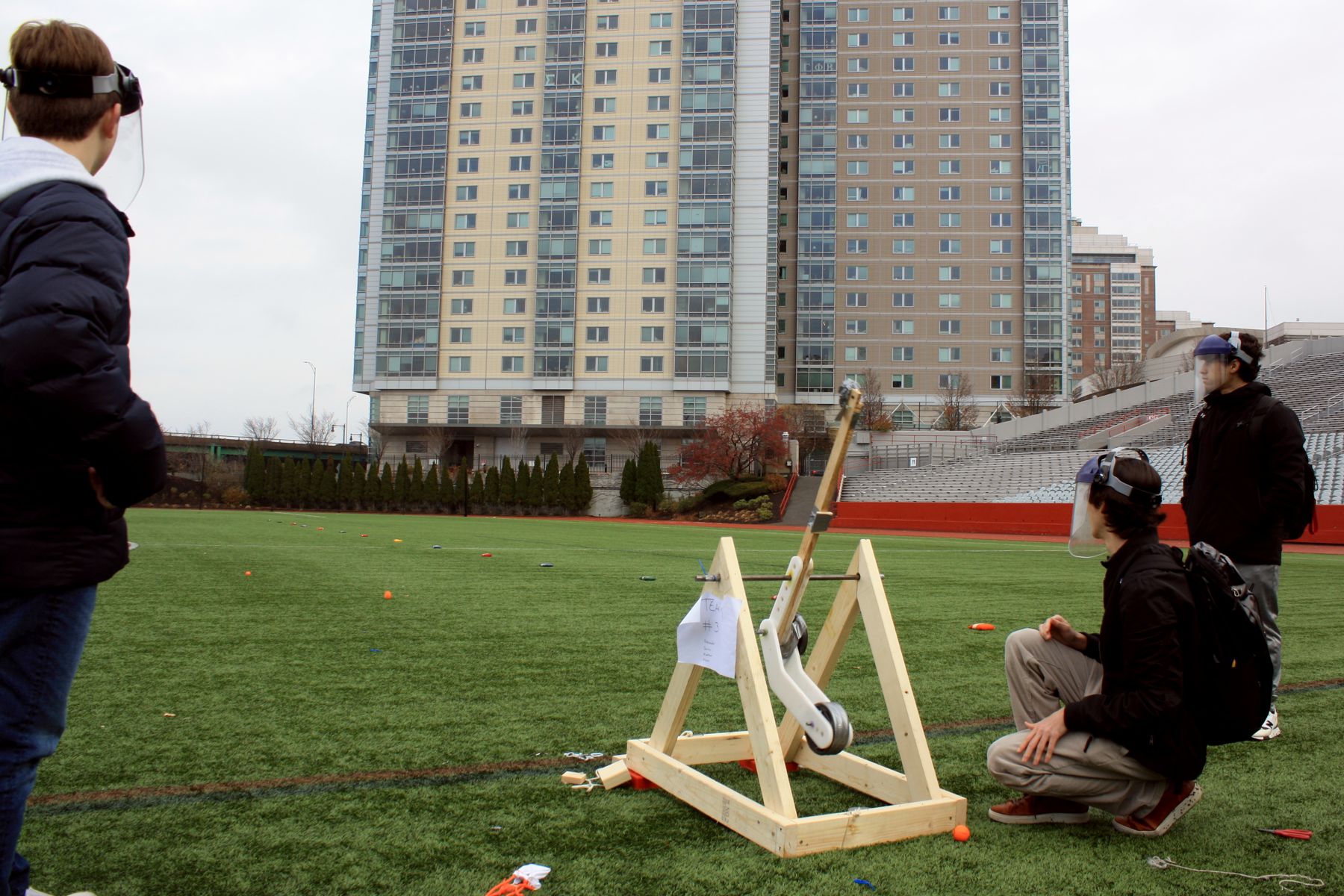long shot of 3 students looking down field at the golf ball just launched by their catapult